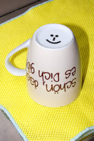 Eine umgedrehte weiße Tasse mit einem Smiley auf dem Boden steht auf einem gelben Stofftuch. An der Seite der Tasse steht ein deutscher Text.