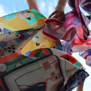 Eine Nahaufnahme aus niedriger Perspektive von bunt gemustertem Stoff, der gehalten wird, mit teilweise sichtbaren Händen und einem blauen Himmel im Hintergrund.