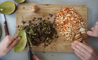 Zwei Hände schneiden Gemüse und Pilze auf einem Holzschneidebrett, außerdem sind ein Messer, eine Schere und eine Avocadoschale zu sehen.