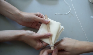 Zwei Paar Hände falten und befestigen weißen Stoff mit einem Holzstab und einer Schnur auf einer grauen Oberfläche.
