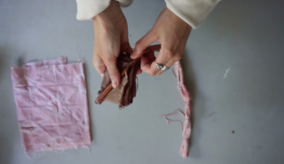 Die Hände einer Person halten braunen Stoff über einem Tisch, auf dem ein gefaltetes rosa Tuch und ein gerollter, zusammengebundener rosa Stoffstreifen daneben liegen.