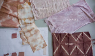 Auf einem Tisch sind verschiedene gefärbte Stoffmuster mit den Aufschriften „Baumwolle“, „Viskose“ und „Seide“ angeordnet, einige davon über einem Papier mit Farbmustern.
