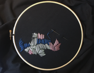 Ein Stickrahmen aus Holz hält schwarzen Stoff mit abstrakten Stichen aus weißem, rosa und blauem Garn; eine mit blauem Garn eingefädelte Nadel liegt auf dem Stoff.