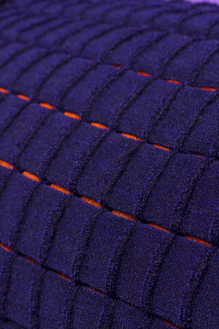Nahaufnahme eines strukturierten violetten Stoffes mit einem erhabenen Gittermuster und dünnen orangefarbenen Linien, die horizontal zwischen den Reihen verlaufen.