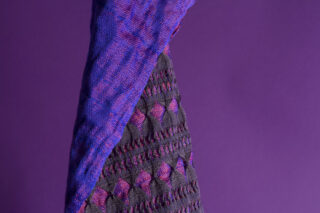 Eine Nahaufnahme eines strukturierten Stoffes mit gewebten Mustern in Schwarz, Lila und Rosa vor einem einfarbigen lila Hintergrund.