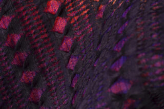 Nahaufnahme eines strukturierten Stoffes mit einem gewebten Muster, bestehend aus schwarzen Fäden auf einem roten und violetten Hintergrund.
