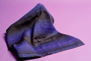Ein blau-schwarz gewebtes Tuch liegt zerknüllt auf einem violetten Untergrund.