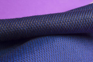 Nahaufnahme eines strukturierten, dunkelblauen Stoffes mit Rautenmuster, der vor einem violetten Hintergrund drapiert ist.