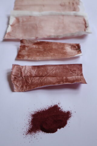 Vier mit rotbraunem Pulver befleckte Papierstreifen sind horizontal über einem kleinen Haufen des gleichen Pulvers auf weißem Hintergrund angeordnet.