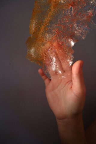 Eine Hand hält ein dünnes, durchscheinendes Blatt mit orangefarbenen und roten Sprenkeln vor einem schlichten Hintergrund hoch.