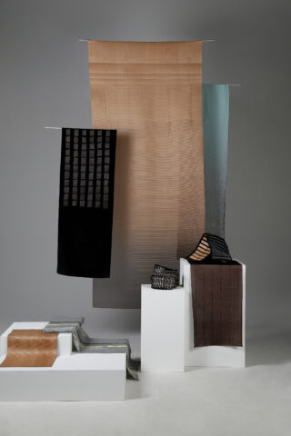 Mehrere Stoffstücke in neutralen und dunklen Tönen sind auf weißen Plattformen drapiert und ausgestellt und hängen in einem minimalistischen Ambiente vor einem grauen Hintergrund.