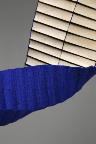Ein gebogenes Objekt mit abwechselnd hellen Holzlatten und einem strukturierten, tiefblauen Stoff, der einen Teil seiner Oberfläche bedeckt, vor einem schlichten grauen Hintergrund.