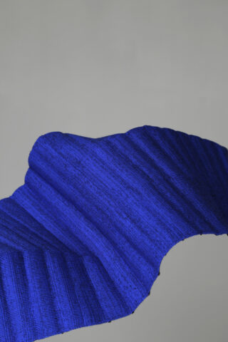 Eine Nahaufnahme eines strukturierten, plissierten Stoffes in einem tiefen Blau, der in einer wellenförmigen Form vor einem schlichten grauen Hintergrund drapiert ist.