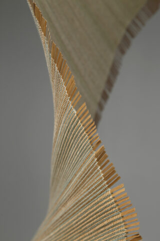 Nahaufnahme eines gewebten Textils mit feinen Goldfäden und ausgefransten Rändern, das vor einem neutralen grauen Hintergrund eine gewundene, bandartige Form bildet.