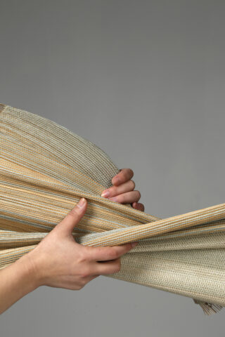 Zwei Hände halten und ziehen ein Stück gewebten beige-gelben Stoff vor einem schlichten grauen Hintergrund auseinander.