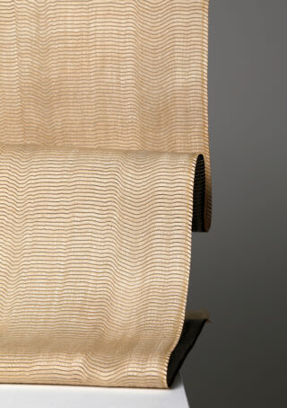 Nahaufnahme eines beigen Stoffes mit einer welligen, horizontal gestreiften Textur, der über eine weiße Oberfläche mit neutralem grauem Hintergrund drapiert ist.