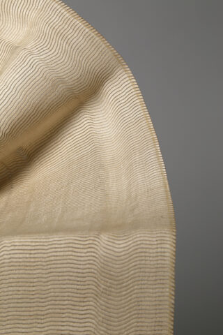 Eine Nahaufnahme eines hellbeigen, halbtransparenten Stoffes mit feinen, gewellten, parallelen schwarzen Linien vor einem schlichten grauen Hintergrund.
