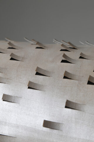 Nahaufnahme einer hellen, strukturierten Oberfläche mit gleichmäßig verteilten rechteckigen Klappen, die ausgeschnitten und teilweise angehoben sind, wodurch ein geometrisches Muster entsteht.