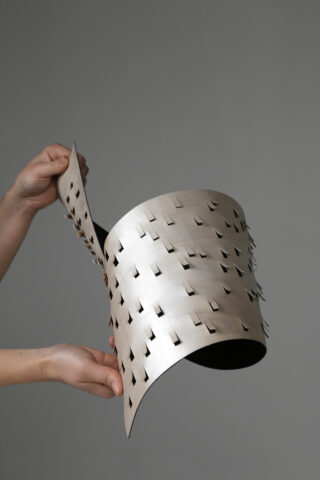 Zwei Hände halten ein flexibles Metallblech mit einem Gitter aus ausgeschnittenen Schlitzen, die vor einem schlichten Hintergrund eine geschwungene, gemusterte Struktur bilden.