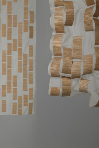 Zwei Stoffbahnen, jede mit einem Muster aus rechteckigen Holzstücken; eine Bahn hängt flach, während die andere geraffte, dreidimensionale Falten aufweist.