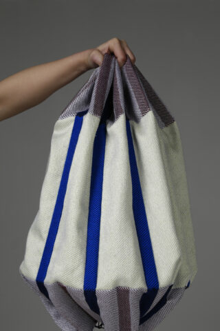 Eine Hand hält eine große gewebte Tasche mit blauen, weißen und braunen vertikalen Streifen auf einem schlichten grauen Hintergrund.