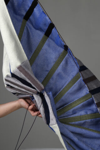 Eine Hand hält ein großes, gefaltetes Textil mit blauen, weißen, grünen und schwarzen geometrischen Streifen vor einem schlichten grauen Hintergrund.