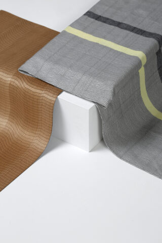 Zwei gemusterte Stoffe, einer braun und einer grau mit einem gelben Streifen, sind über einem weißen rechteckigen Block auf einer weißen Oberfläche drapiert.