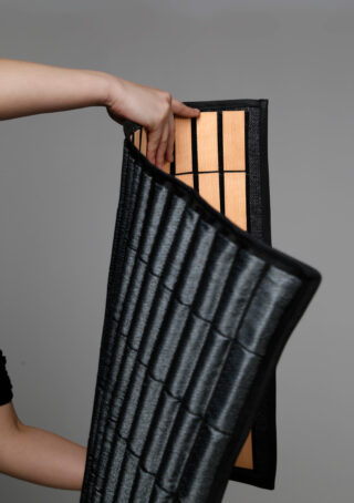 Eine Hand hält eine faltbare Matte mit einer schwarzen Polsterung außen und einem Abschnitt mit freiliegenden Bambuslatten innen.