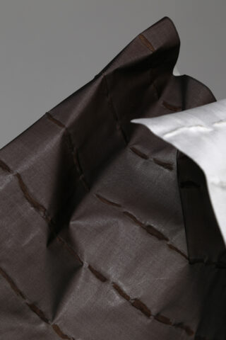 Eine Nahaufnahme von zwei Stoffstücken, eines dunkelbraun mit sichtbaren Nähten und eines hellgrau, die in überlappenden, eckigen Falten angeordnet sind.