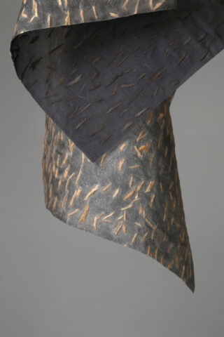 Eine Nahaufnahme eines strukturierten, gefalteten Stücks dunklen Papiers mit hellbraunen, pinselstrichartigen Mustern auf einem schlichten grauen Hintergrund.