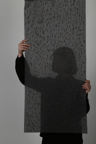 Eine Person hält ein halbtransparentes Netztuch mit kleinen, dunklen Formen darauf hoch, die ihr Gesicht und ihren Oberkörper verdecken.
