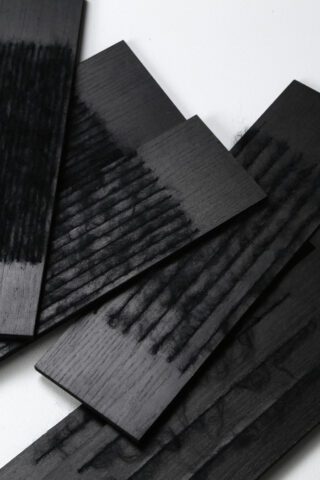 Mehrere rechteckige Stücke aus schwarzem Holz mit sichtbaren Strukturmustern sind in überlappenden Schichten auf einer weißen Oberfläche angeordnet.