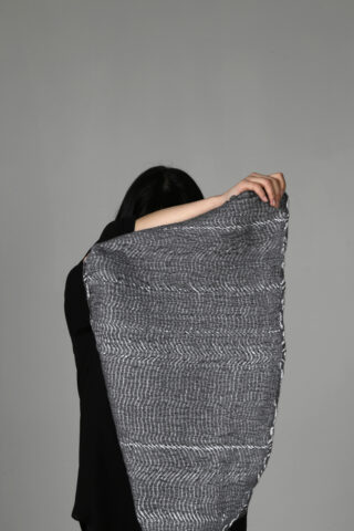 Eine Person hält vor einem schlichten grauen Hintergrund ein großes, strukturiertes graues Tuch vor ihr Gesicht.