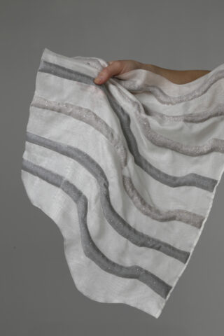 Eine Hand hält einen weißen Stoff mit wellenförmigen, parallelen grauen und silbernen Strukturstreifen vor einem schlichten grauen Hintergrund.