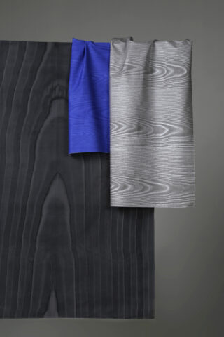 Drei Stoffstücke mit Holzmaserung in Schwarz, Blau und Silber sind vor einem schlichten grauen Hintergrund drapiert.