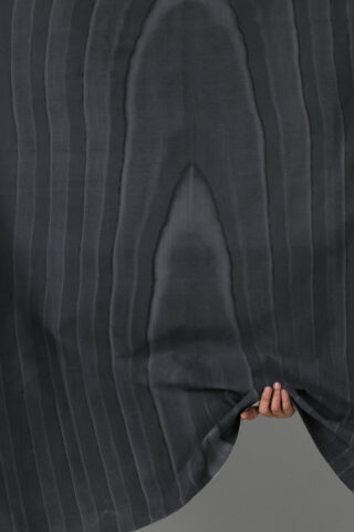 Eine Hand hält die untere Ecke eines dunklen, vertikal gestreiften Stoffes mit einem subtilen, organischen Wellenmuster.