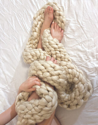 Zwei Füße und zwei Hände sind teilweise in dickes, klobiges cremefarbenes Garn eingewickelt und liegen auf einem weißen Laken.