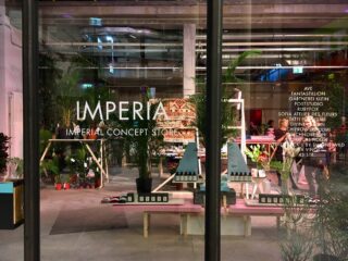 Eine Glasfassade mit der Aufschrift „IMPERIAL CONCEPT STORE“ gibt den Blick auf ein modernes Interieur mit Pflanzen, farbenfrohem Dekor und verschiedenen Schaufenstern frei.