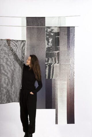 Eine schwarz gekleidete Frau steht vor abstrakten, transparenten Textilien mit wellenförmigen, geometrischen Mustern und hält einen Stoff mit ihrer rechten Hand hoch.