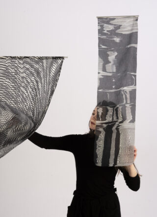 Eine Person in schwarzer Kleidung hält zwei Stücke halbtransparenten, gemusterten Stoff hoch; eines ist horizontal und gewellt, das andere vertikal mit unregelmäßigen Mustern.