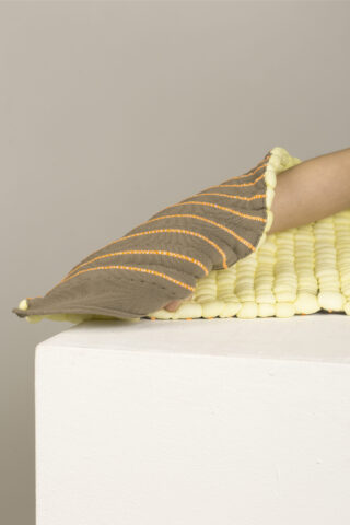 Eine Hand mit einem strukturierten, gestreiften Handschuh berührt eine Oberfläche, die mit kleinen, weichen, gelben Schaumstoffperlen auf einem weißen Sockel bedeckt ist.