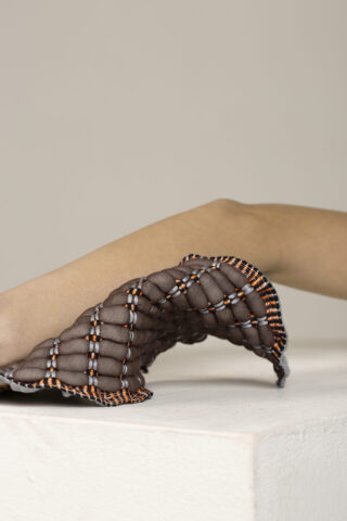 Der Arm einer Person ruht auf einer weißen Oberfläche, die teilweise mit einem braunen, gesteppten und strukturierten Stoff mit orangefarbenen und weißen Nähten bedeckt ist.