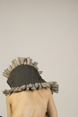 Eine Person mit nacktem Rücken trägt vor einem schlichten hellen Hintergrund ein strukturiertes, graues Kopfstück, das mit beigen, gerollten Stoffdetails verziert ist.