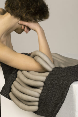 Eine Person sitzt da, der Kopf ruht auf der Hand, der Arm liegt über einer Oberfläche, die mit weichem, gewickeltem, schlauchartigem Stoff bedeckt ist.