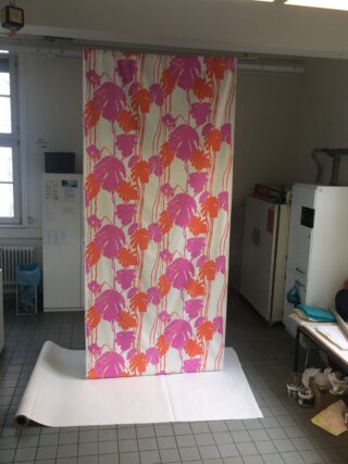 Ein großes Stück Stoff mit einem rosa-orangen Palmenmuster hängt vertikal in einem gefliesten Raum mit Kunstbedarf und einem Fenster.
