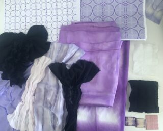 Eine Sammlung violetter, weißer und schwarzer Stoffmuster und -proben, die auf einer flachen Oberfläche angeordnet sind, neben gedruckten geometrischen Musterblättern in passenden Farben.