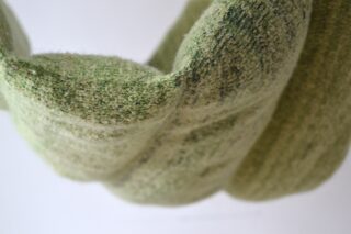 Nahaufnahme eines strukturierten, hellgrünen Strick- oder Webstoffs mit sichtbaren Fasern und einer gefalteten, wellenförmigen Form vor einem schlichten Hintergrund.