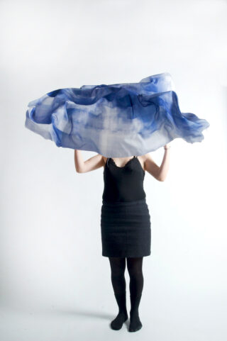 Eine Person in einem schwarzen Outfit hält vor einem schlichten weißen Hintergrund einen großen blau-weißen durchscheinenden Stoff hoch, der ihren Kopf und Oberkörper teilweise verdeckt.