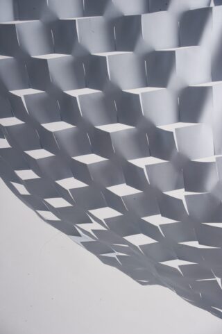 Eine Nahaufnahme eines verwobenen Gittermusters aus weißen Stoffstreifen, das eine geometrische, dreidimensionale Oberfläche mit schrägen Schatten erzeugt.