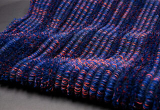 Nahaufnahme einer strukturierten Oberfläche mit dicht gepackten, gewundenen Fasern in Blau- und Rottönen, die in parallelen Reihen auf einem dunklen Hintergrund angeordnet sind.